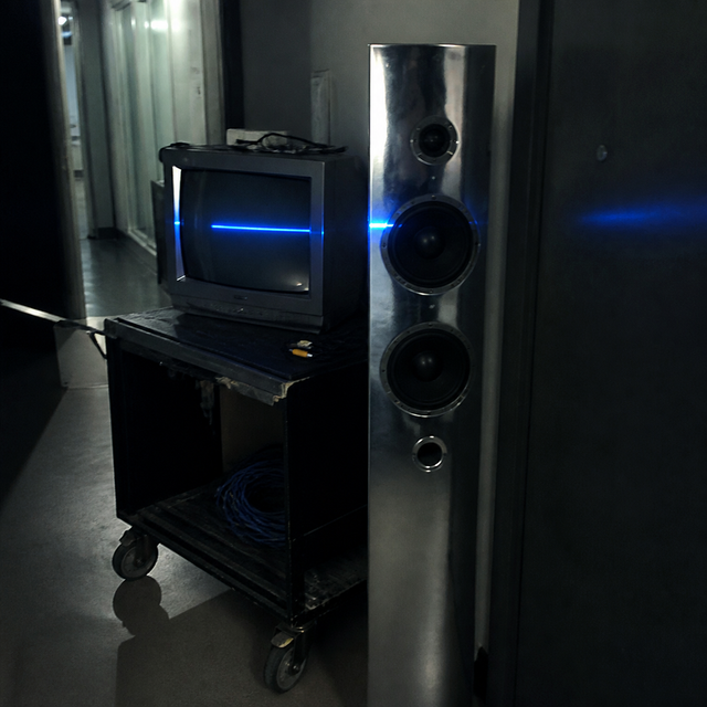 A chrome speaker and dusty CRT monitor sit on an equipment cart in a studio hallway with a cobalt reflection.